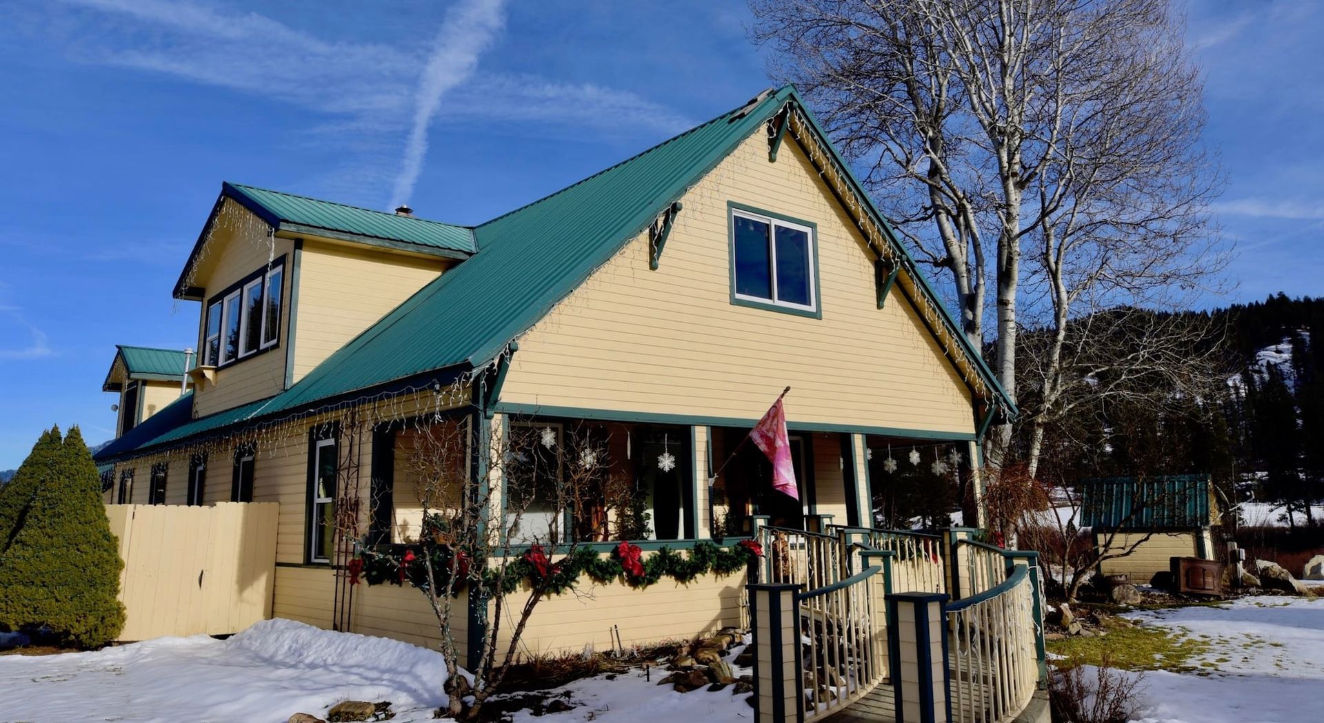 A yellow house with a green roof and a porch decorated for christmas