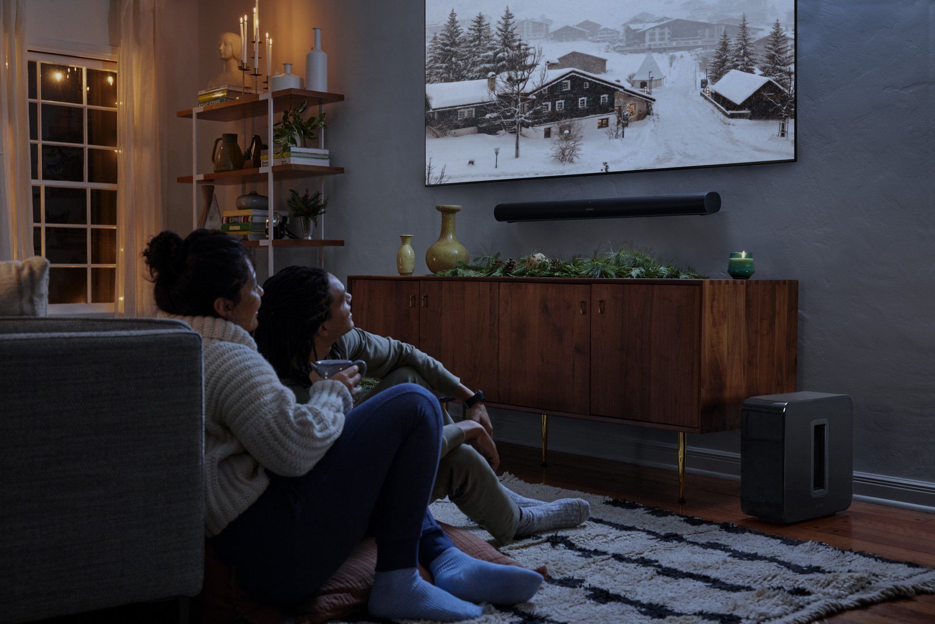 Two people watching a winter scene on TV in a cozy living room, low lighting, sound bar below.