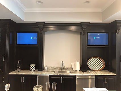Two TVs mounted in a dark-wood bar area with a sink, cabinets, and granite countertops.