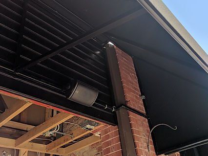 Black speaker attached to a brick pillar under a black awning with a metal frame.