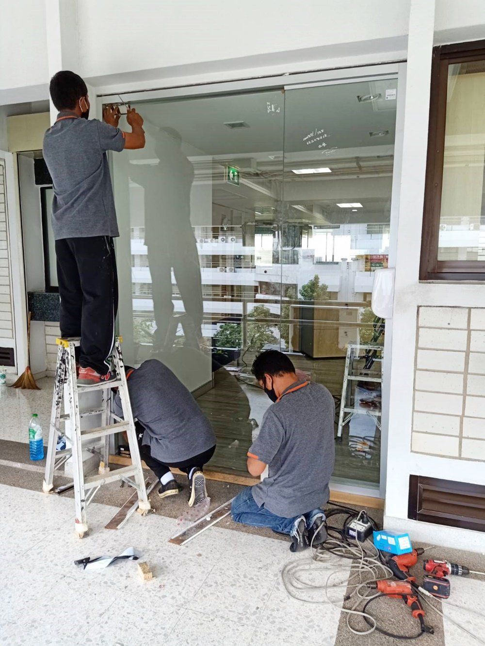 Tres trabajadores instalando una gran puerta de cristal en un edificio. Uno subido a una escalera, dos en el suelo.