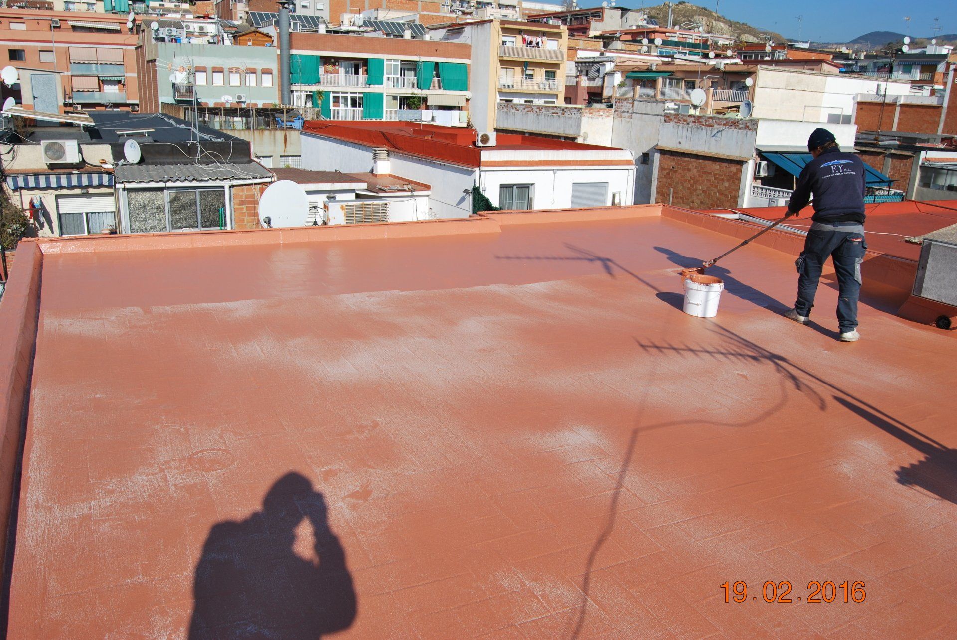 Techador pintando una capa de pintura roja sobre una azotea plana con edificios de la ciudad al fondo en un día soleado.