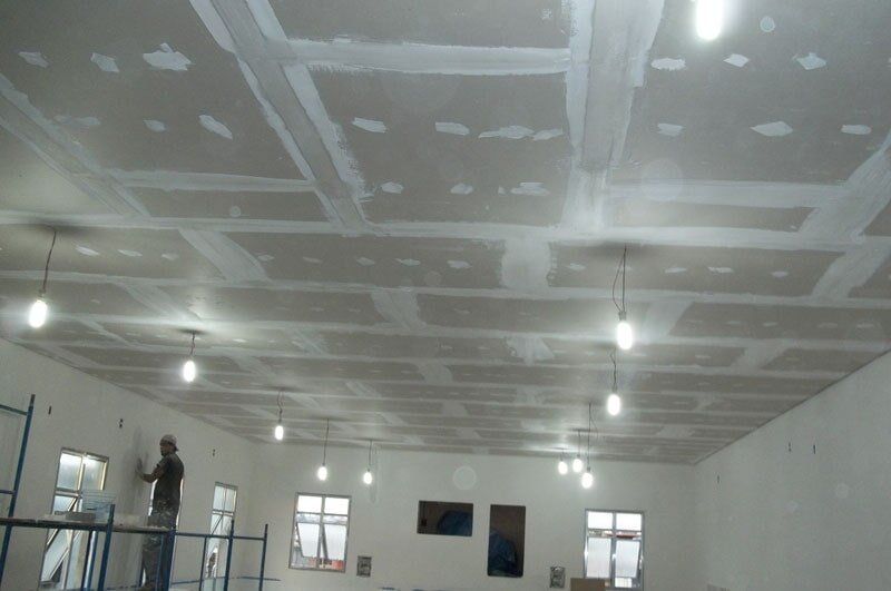 Un hombre subido a un andamio está encintando el techo de pladur; las luces del techo iluminan la habitación.