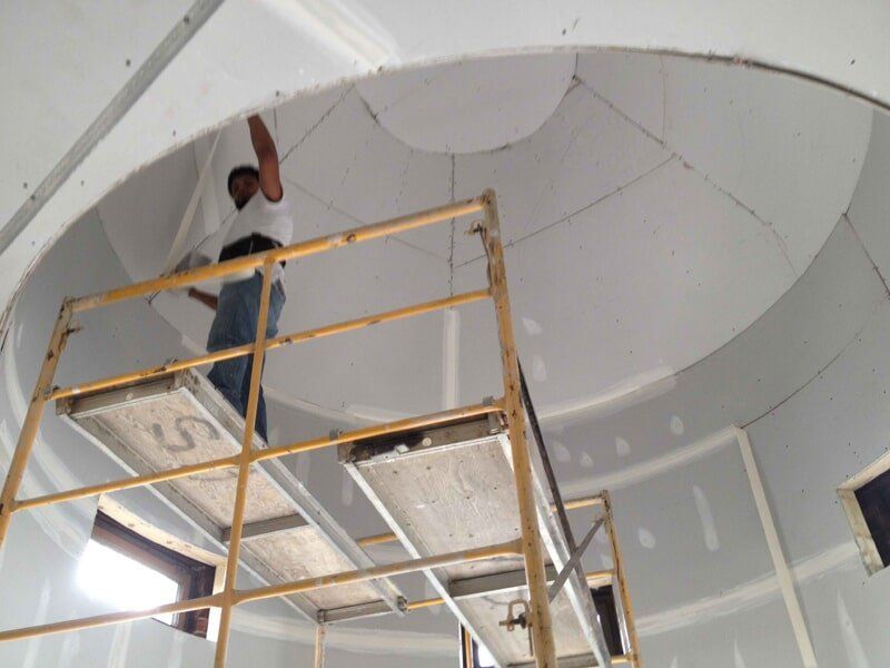 Persona sobre un andamio instalando paneles de yeso en un techo curvo.