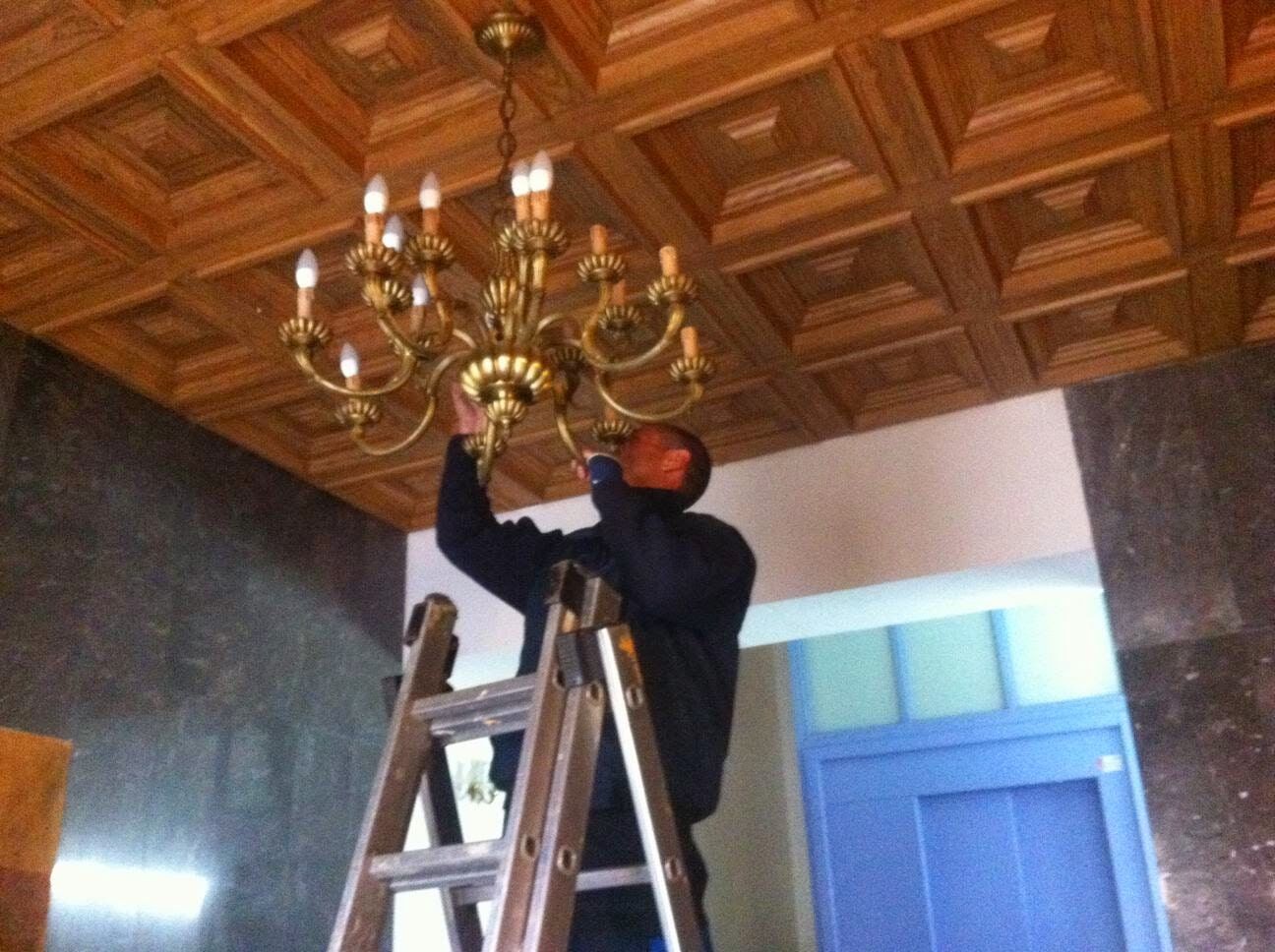 Hombre subido a una escalera arreglando una lámpara de araña dorada en un techo de madera.