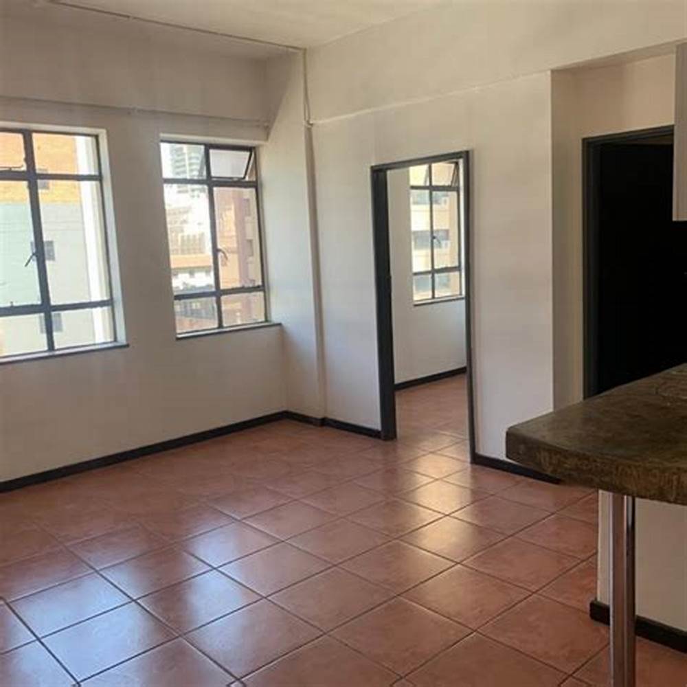 Habitación vacía con suelo de baldosas, dos ventanas y una puerta abierta a otra habitación.