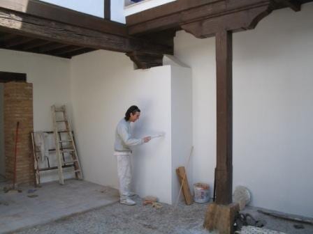 Hombre pintando una pared blanca en un patio parcialmente cubierto. Se ven vigas de madera y una escalera.