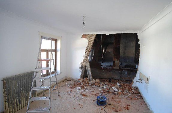 Habitación con una pared parcialmente derribada; escombros en el suelo, escalera junto a la ventana, radiadores.