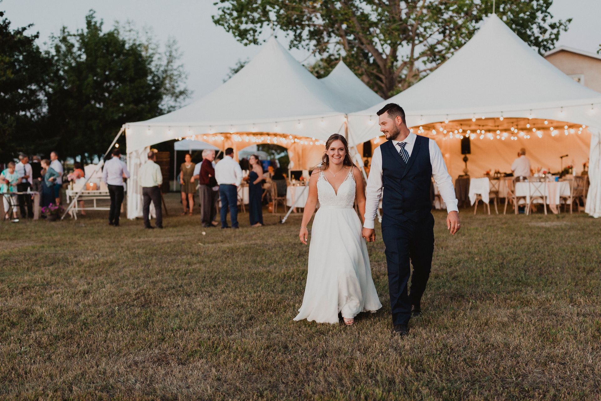 A bride and groom are walking in front of a tent at their wedding reception.