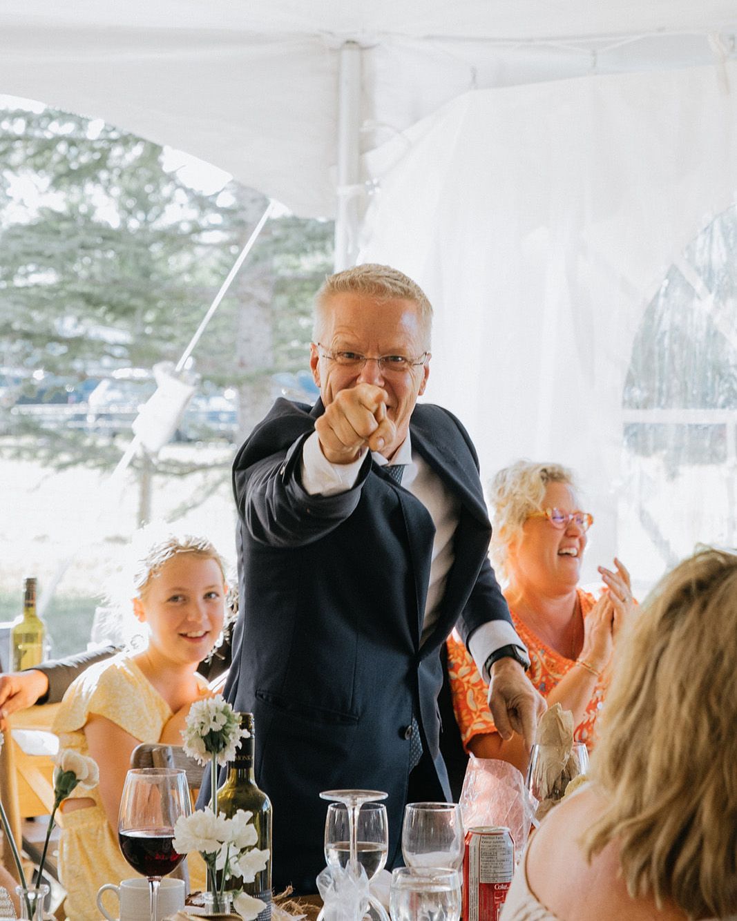 A man in a suit is pointing at the camera at a wedding reception.