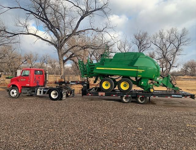 Red truck towing a green John Deere hay baler on a trailer, parked on gravel.