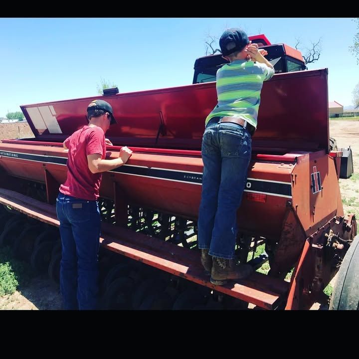 Two people standing near a red agricultural machine under a blue sky, checking or filling it.