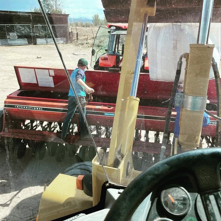 Person loading seeds into a red farm implement, another tractor in the background, view from tractor cab.