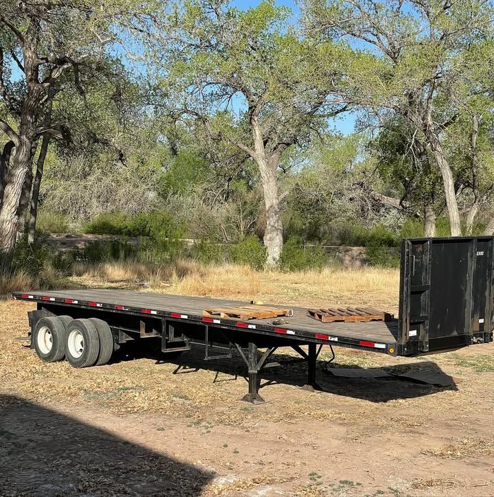 Flatbed trailer with black frame and rear container, parked outdoors on dirt.