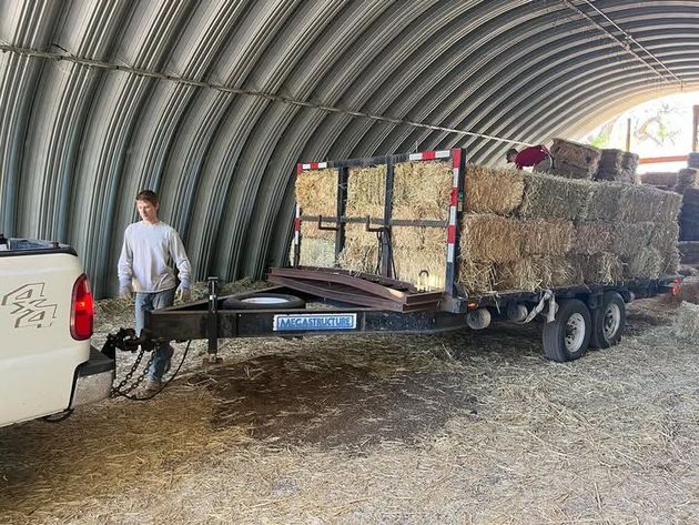 A person stands by a truck and trailer loaded with hay bales inside a metal arched structure.