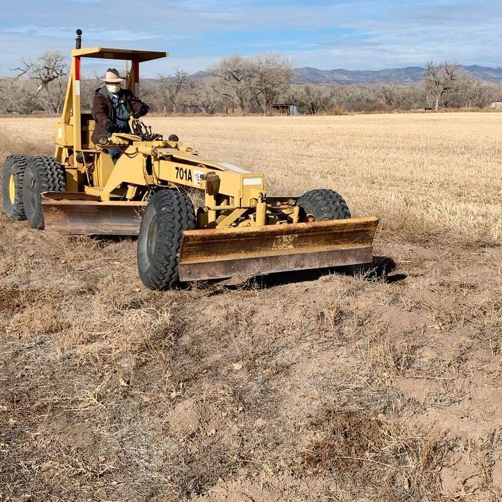 A person operates a yellow grader on a field; mountains in the background.