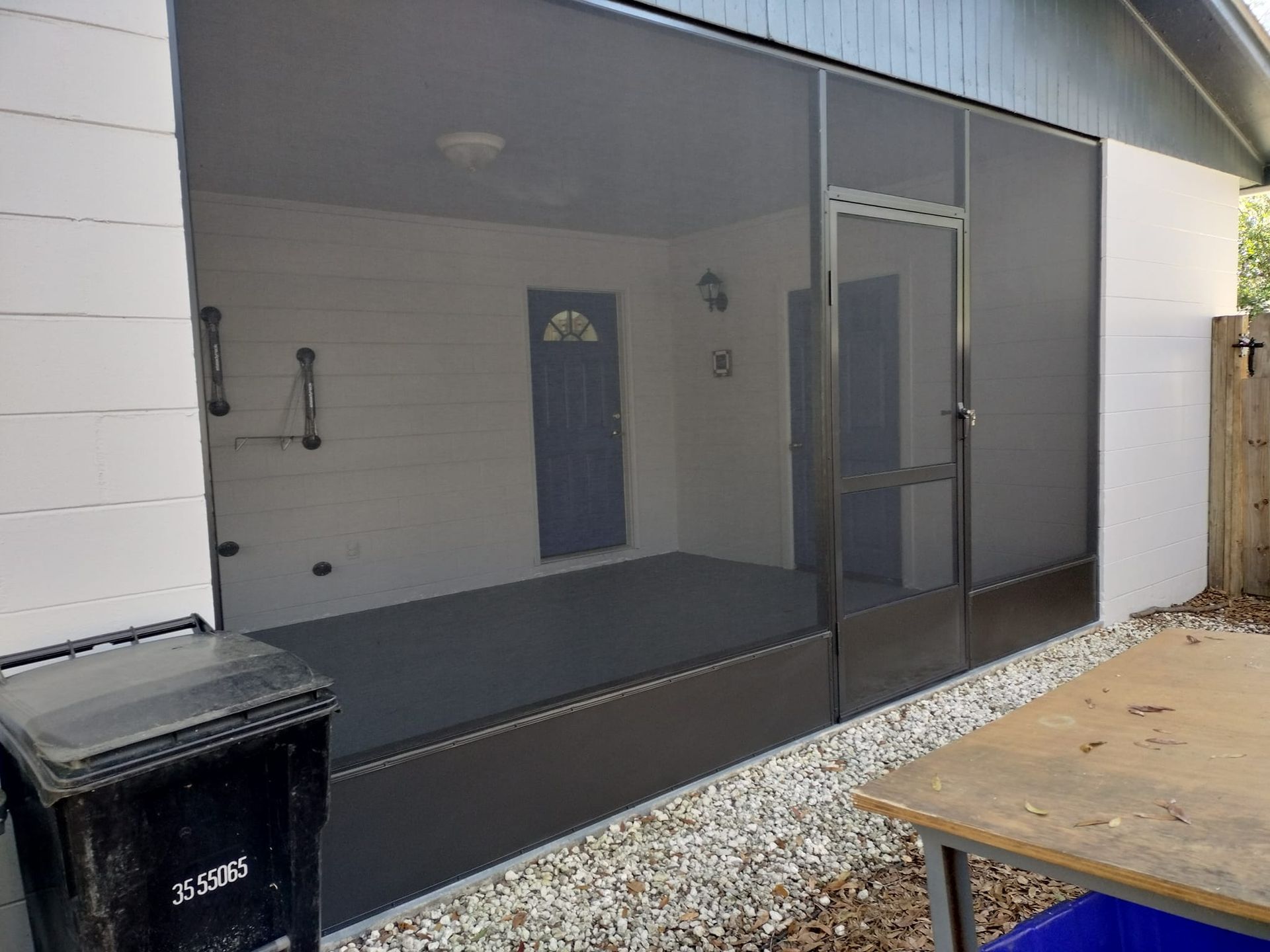 A screened in porch with a black trash can in front of it.