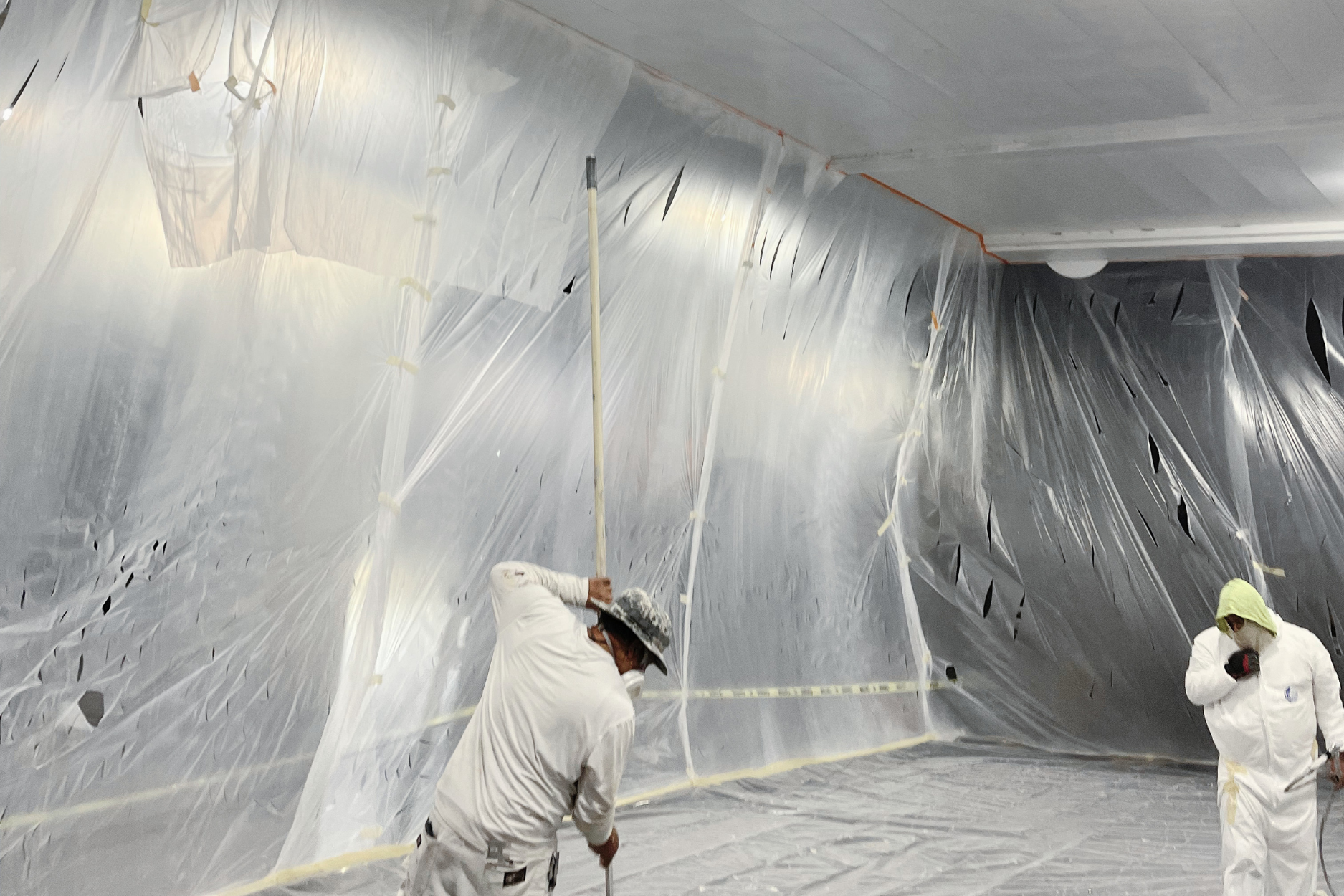 A construction worker in a white uniform kneels on the floor of a home renovation site filled with supplies and trash bags.
