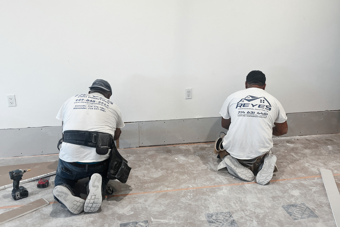 Two workers in white t-shirts kneel on the floor while installing drywall panels along the base of a white wall.