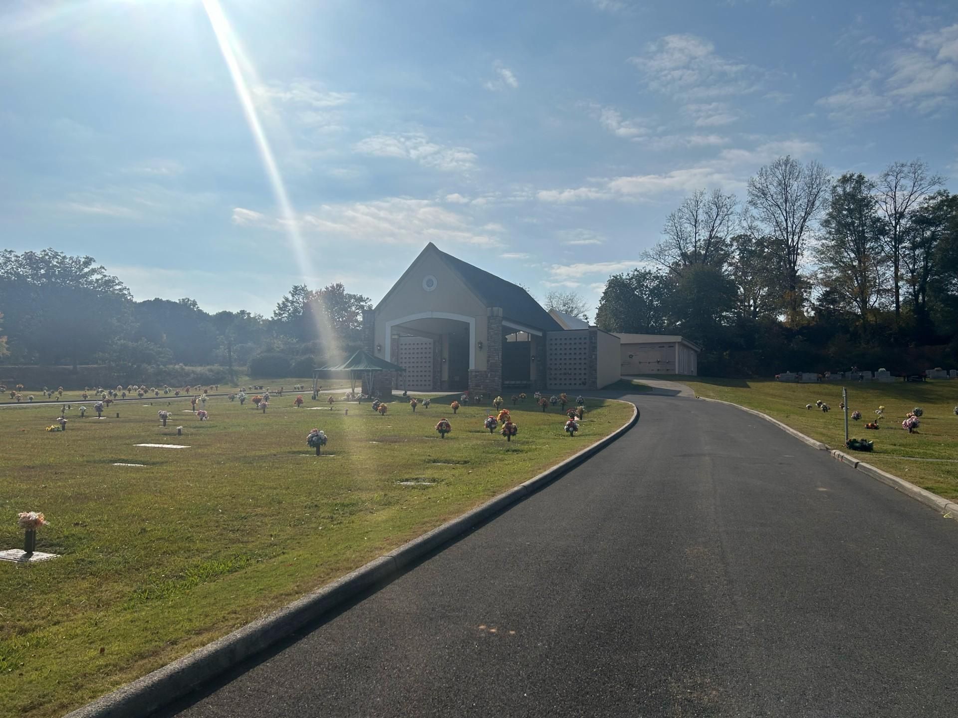 Asphalt road leads to a chapel in a cemetery with headstones and trees under a sunny sky.