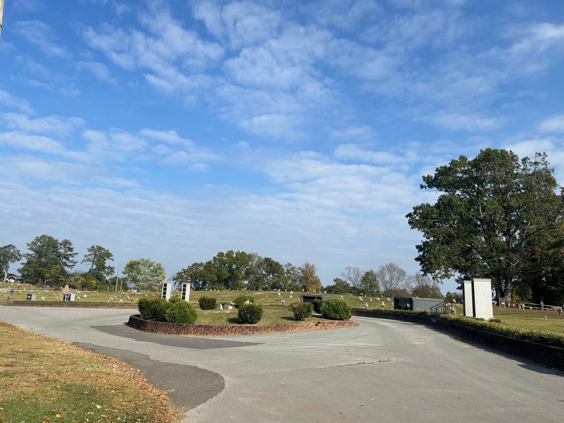 Cemetery with a circular drive, headstones, and trees under a cloudy blue sky.
