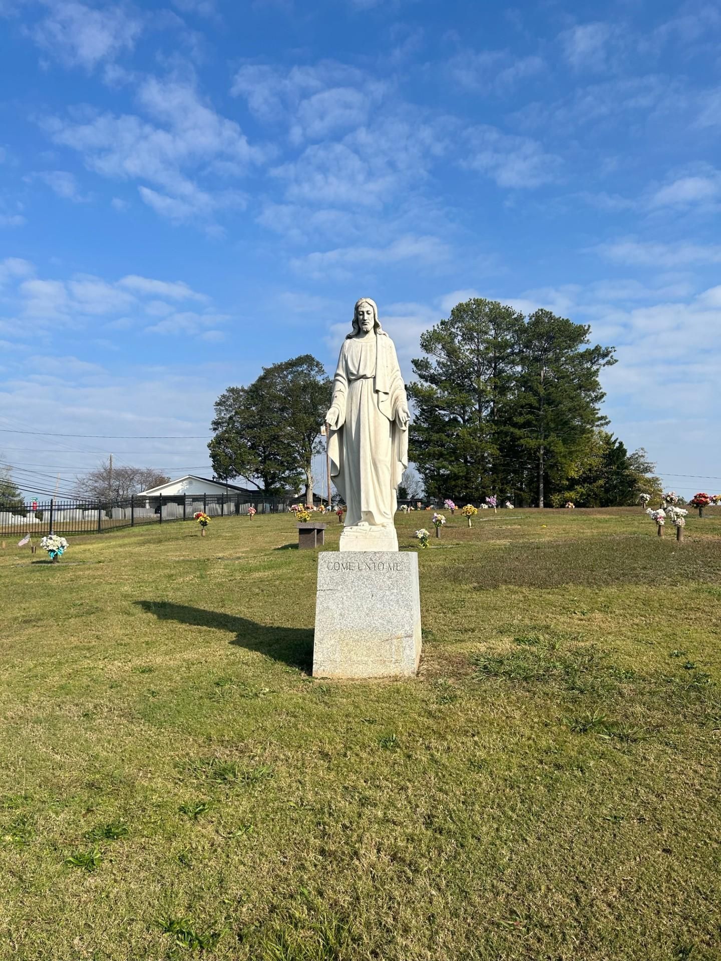 Statue of a robed figure stands on a pedestal in a cemetery; trees and blue sky are in the background.