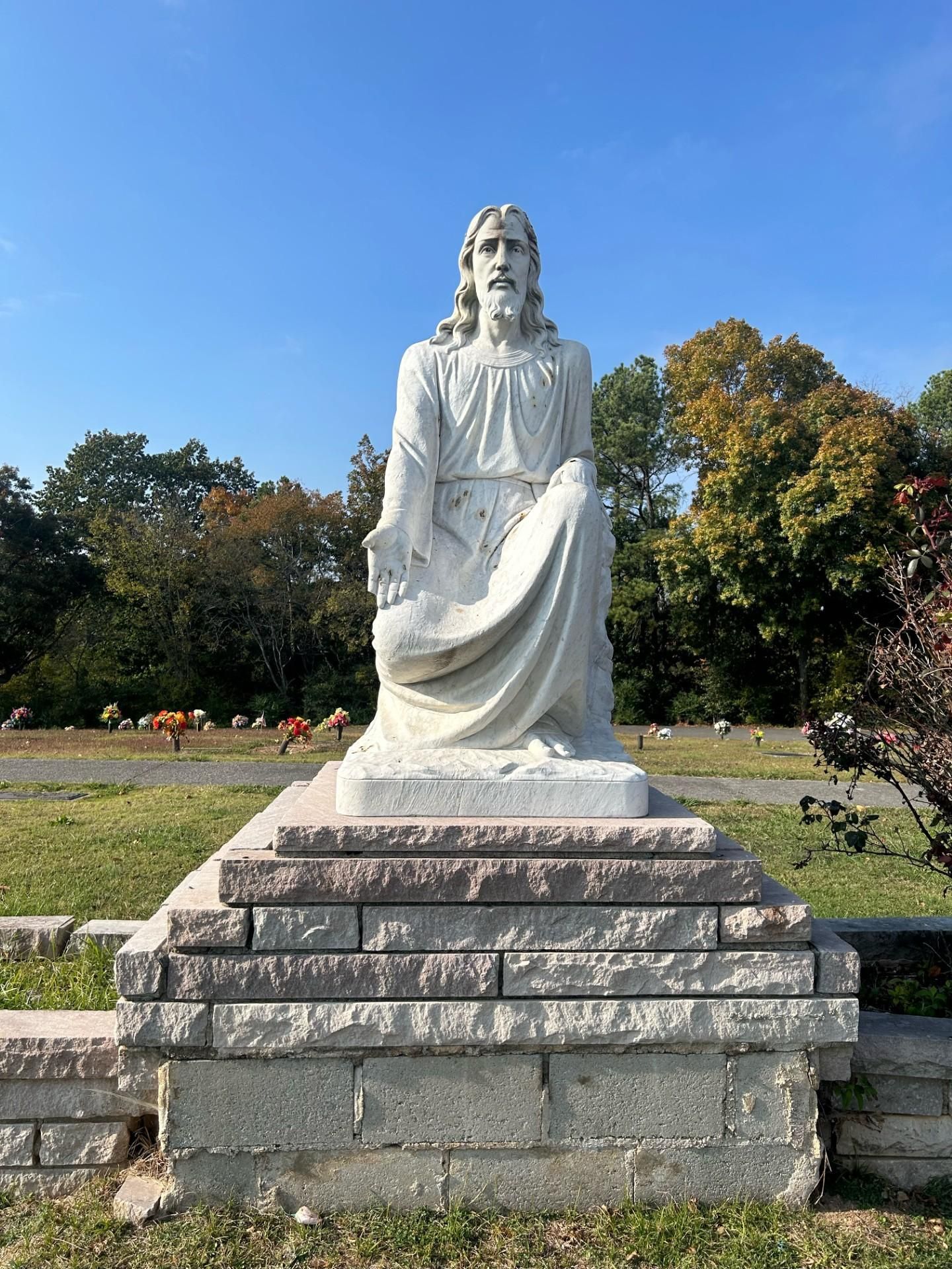 White statue of Jesus kneeling, set on a stone platform, in a cemetery on a sunny day.