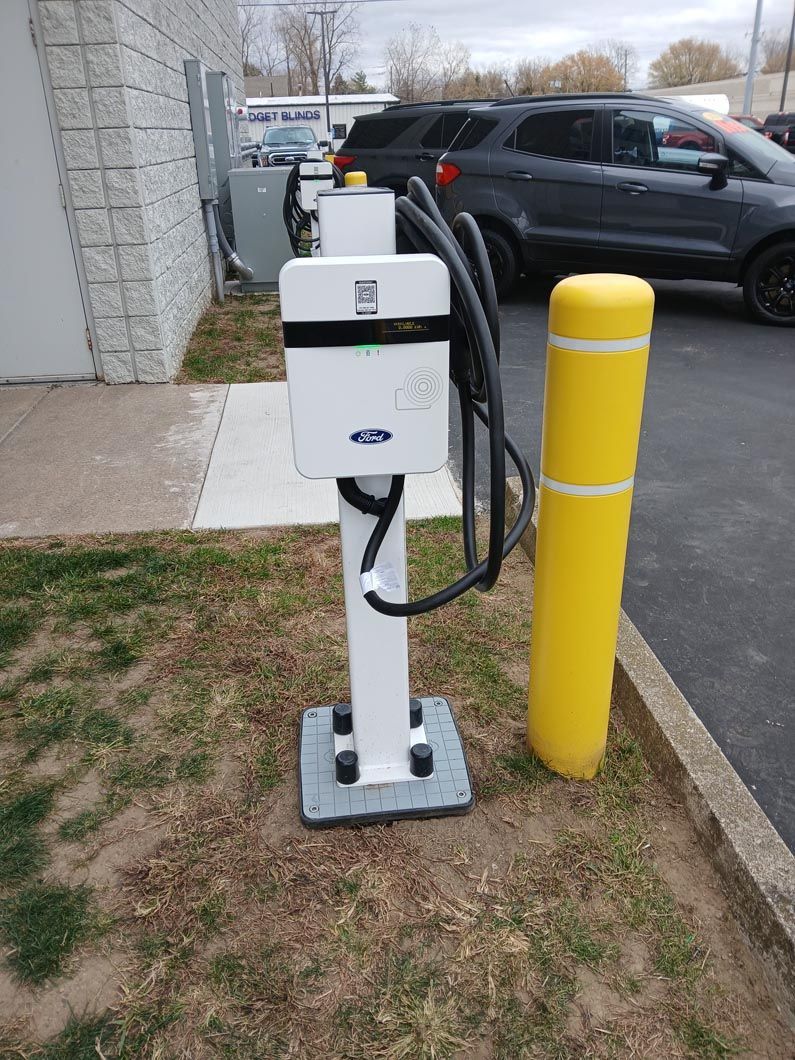 A white charging station is sitting next to a yellow pole in a parking lot.