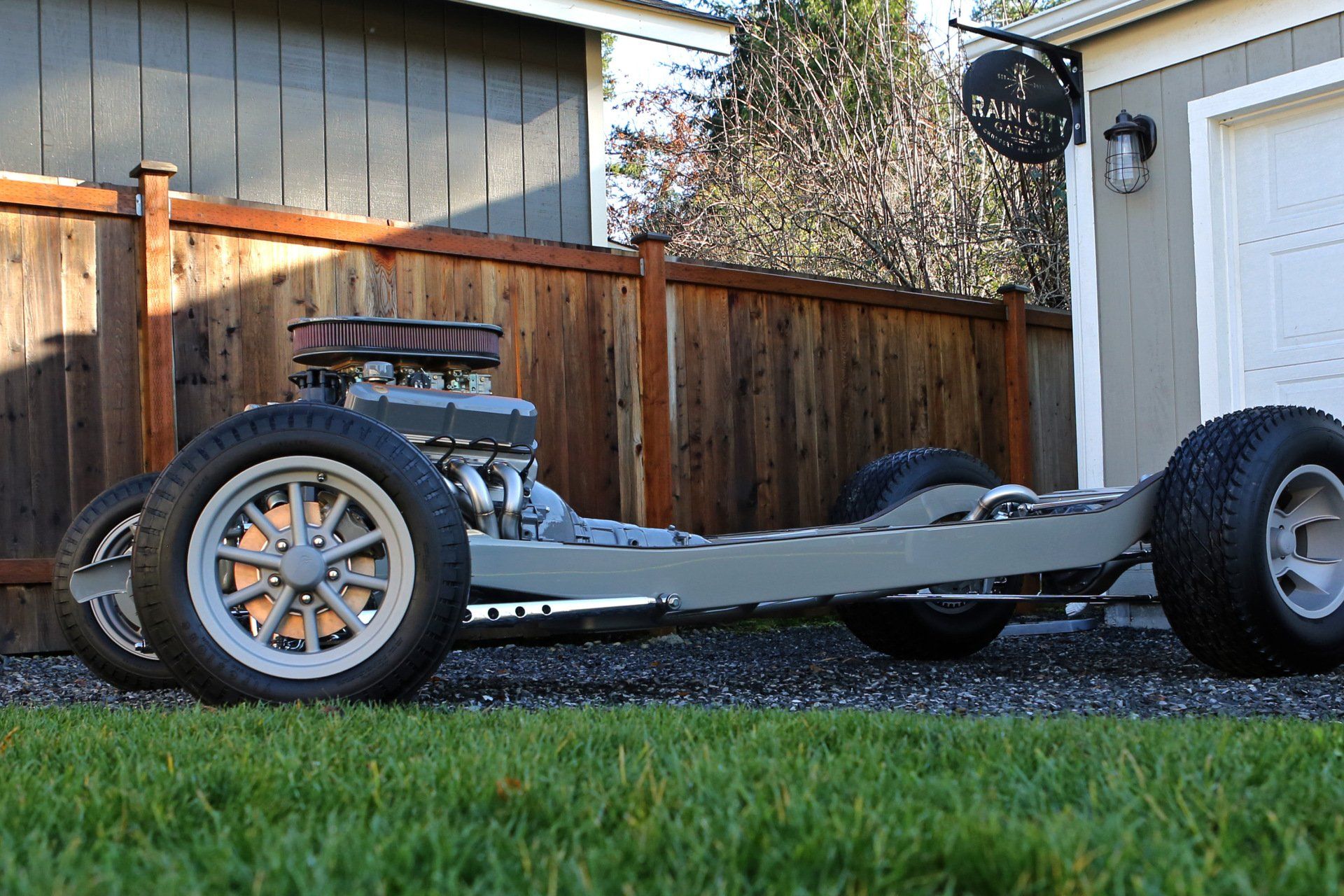 A car frame is sitting in the grass in front of a garage.