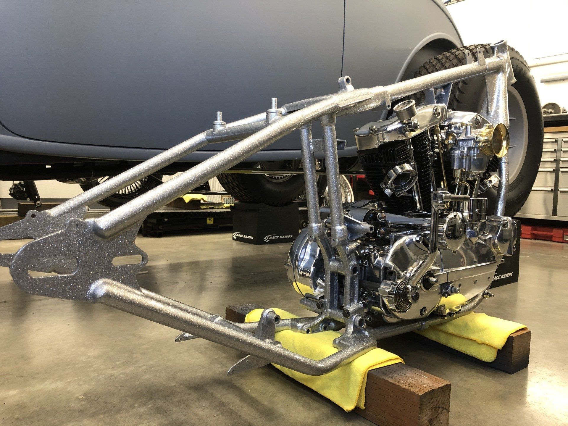 A motorcycle frame is sitting on a wooden block next to a car.