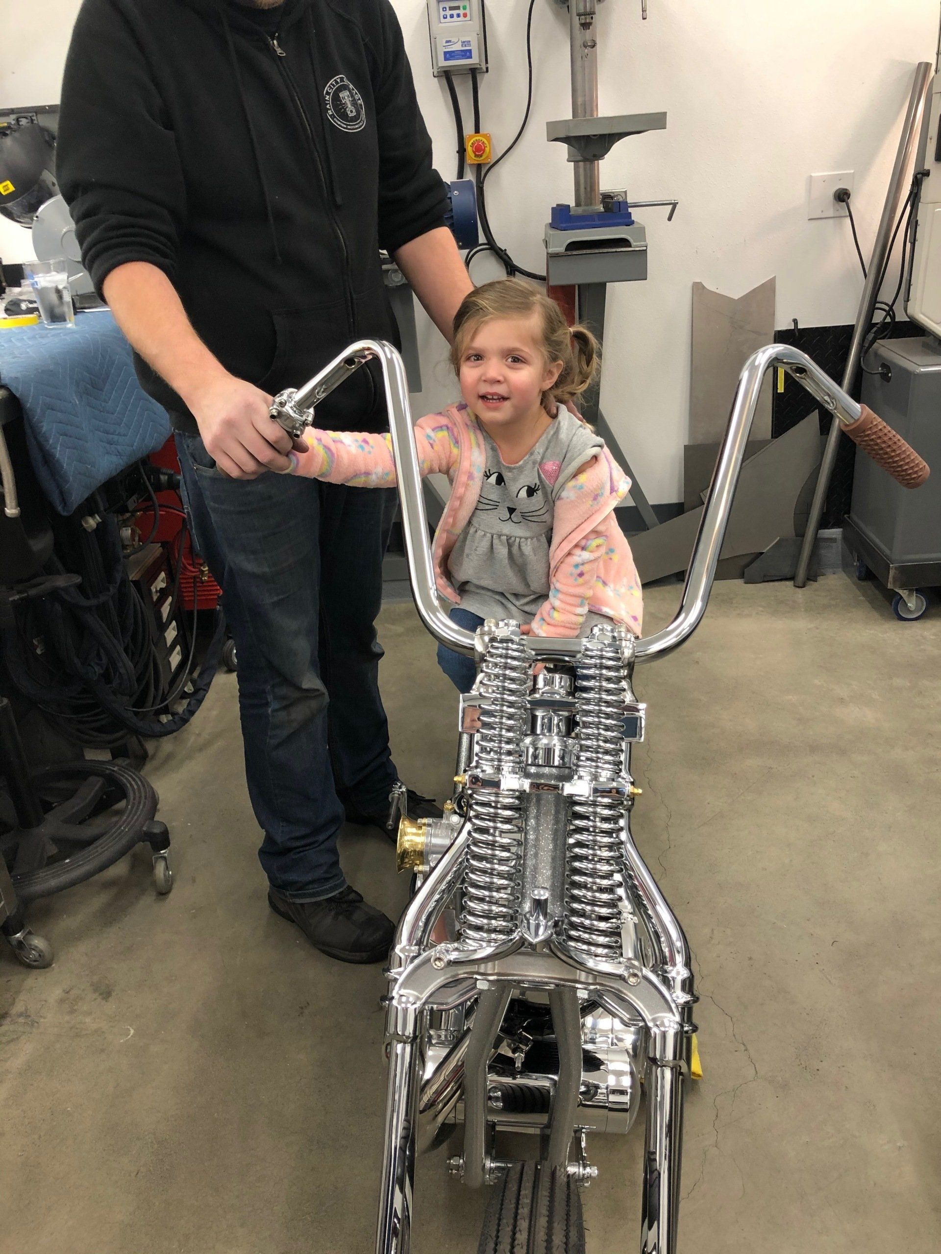 A little girl is sitting on a motorcycle with a man standing behind her.