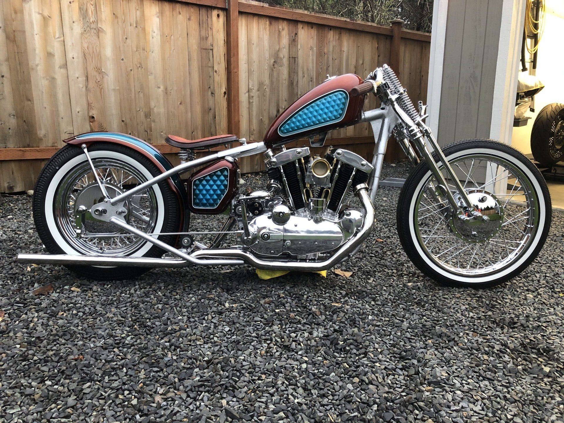 A chopper motorcycle is parked on gravel in front of a wooden fence.
