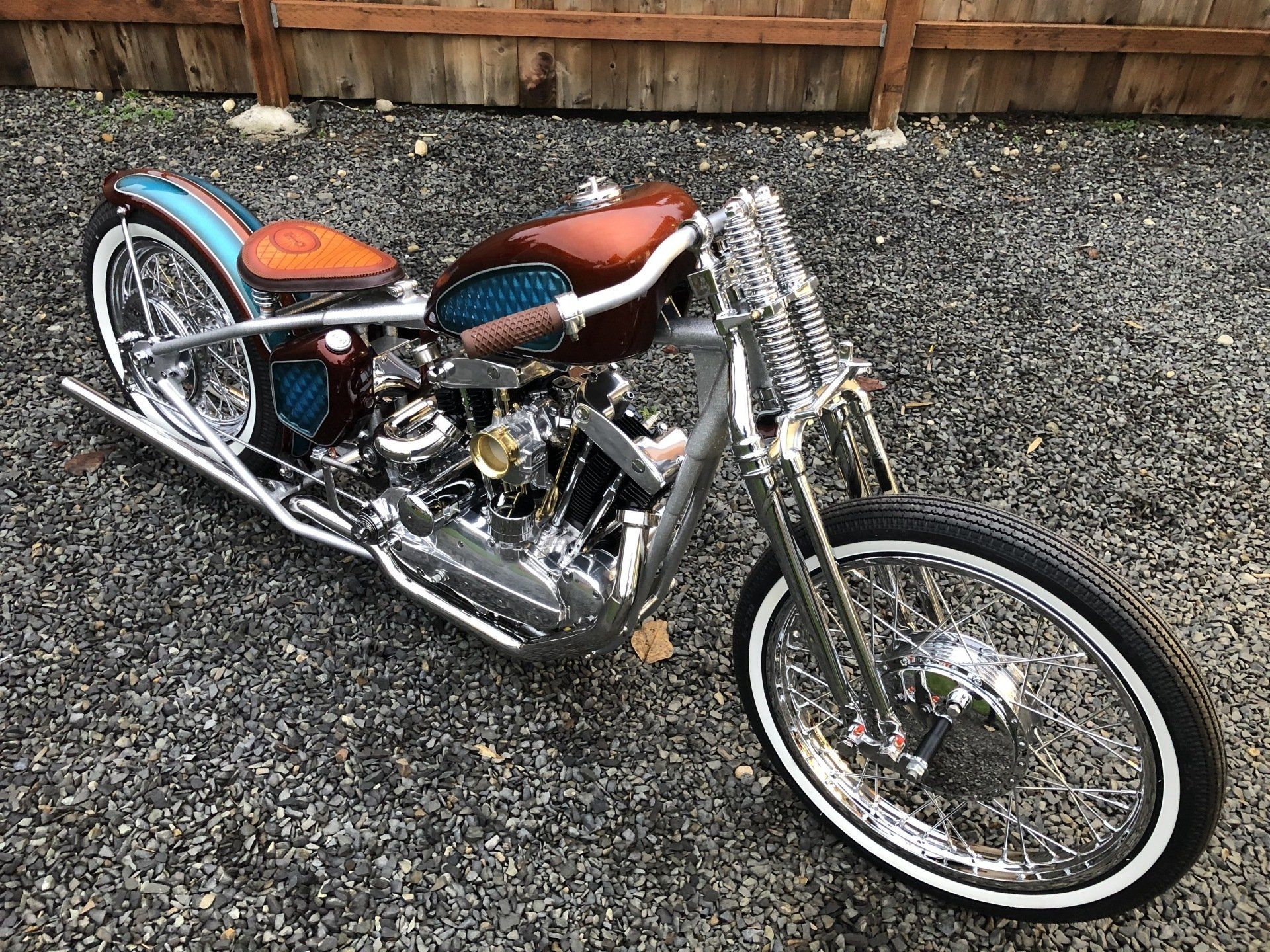 A chopper motorcycle is parked on gravel in front of a wooden fence.