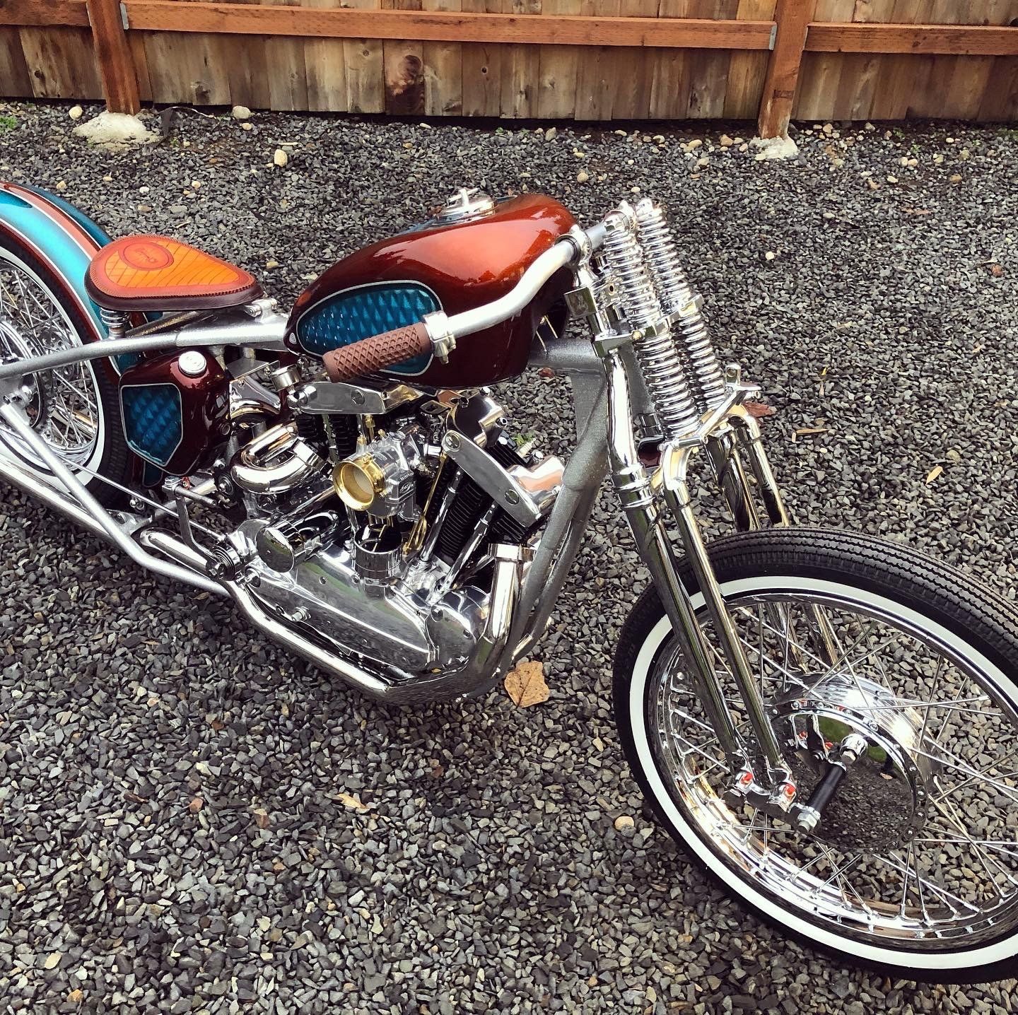 A chopper motorcycle is parked on gravel in front of a wooden fence.