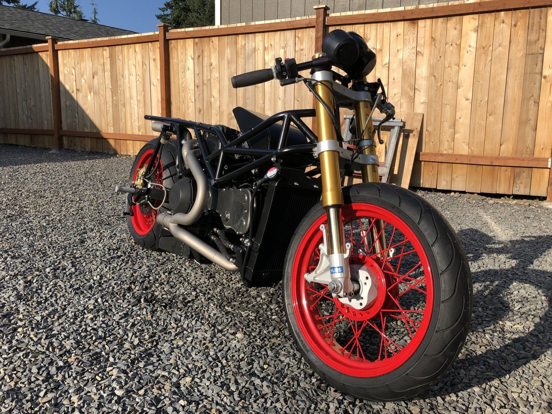 A black motorcycle with red wheels is parked in a gravel lot in front of a wooden fence.