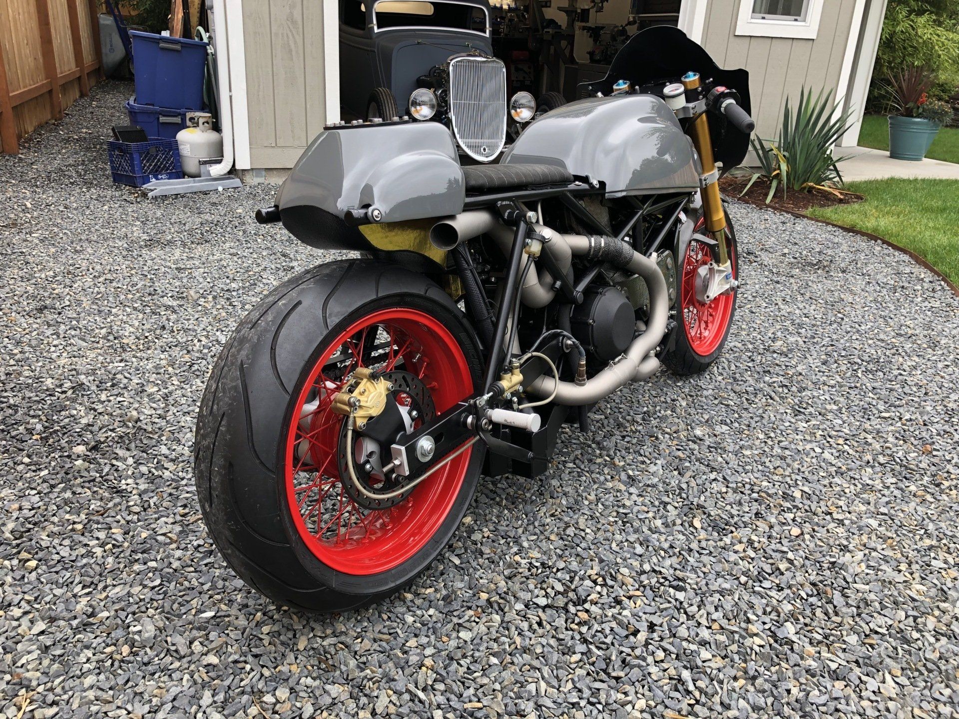 A motorcycle with red wheels is parked on gravel in front of a garage.