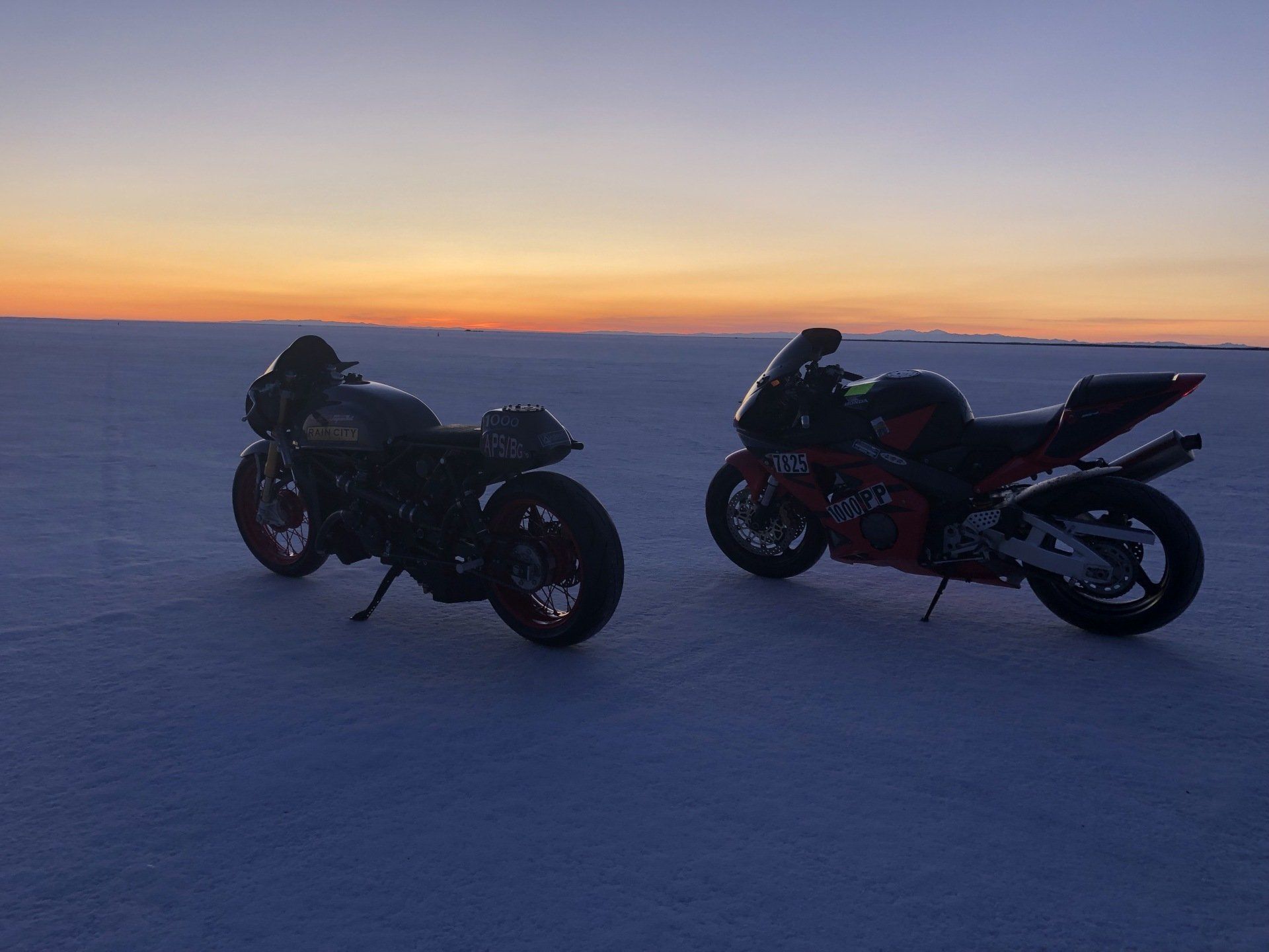 Two motorcycles are parked in the snow at sunset.