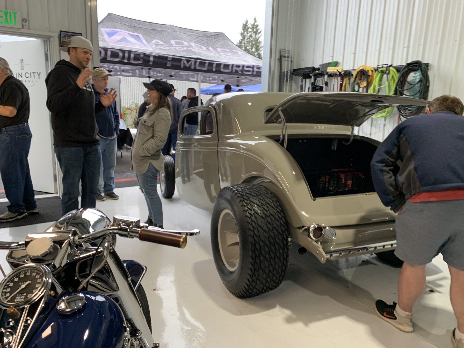 A group of people are standing around a car and a motorcycle in a garage.