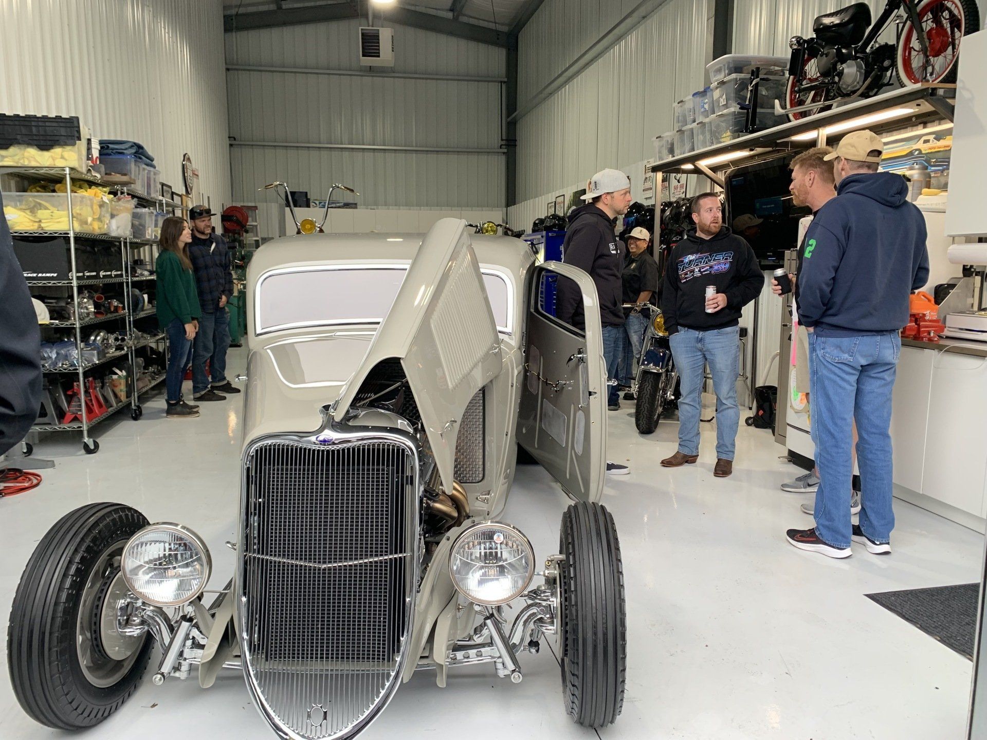 A group of people are standing around a hot rod in a garage.