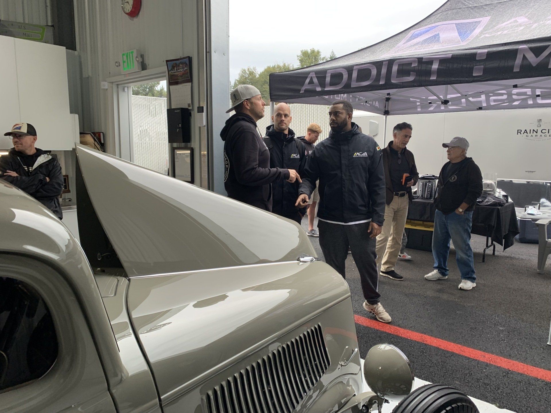 A group of men are standing around a car under a tent that says addict.