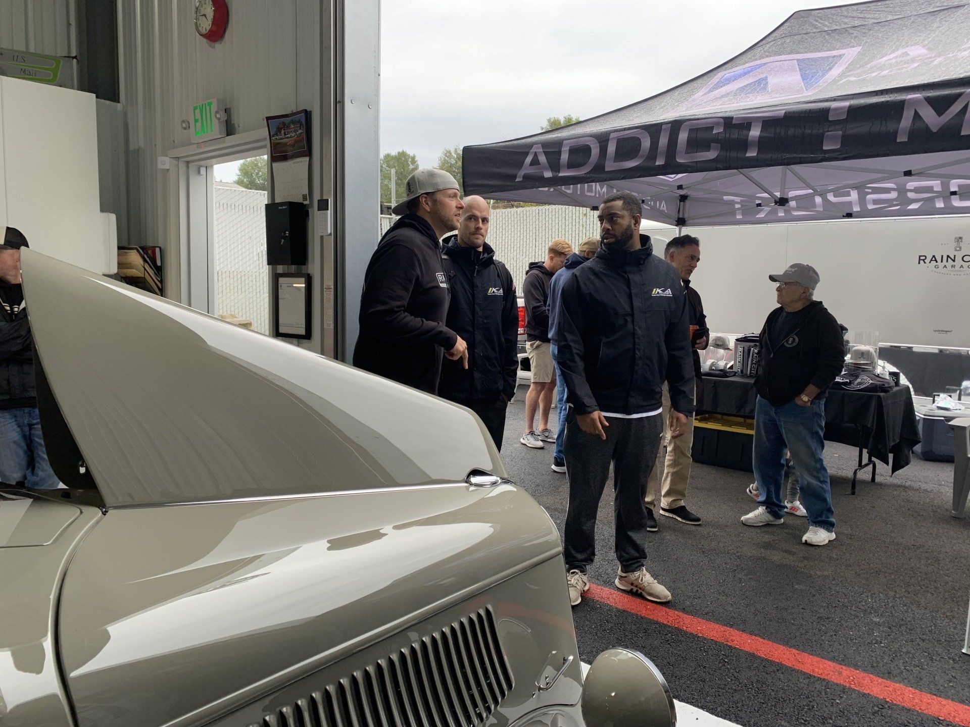 A group of men are standing in front of a car under a tent that says addict