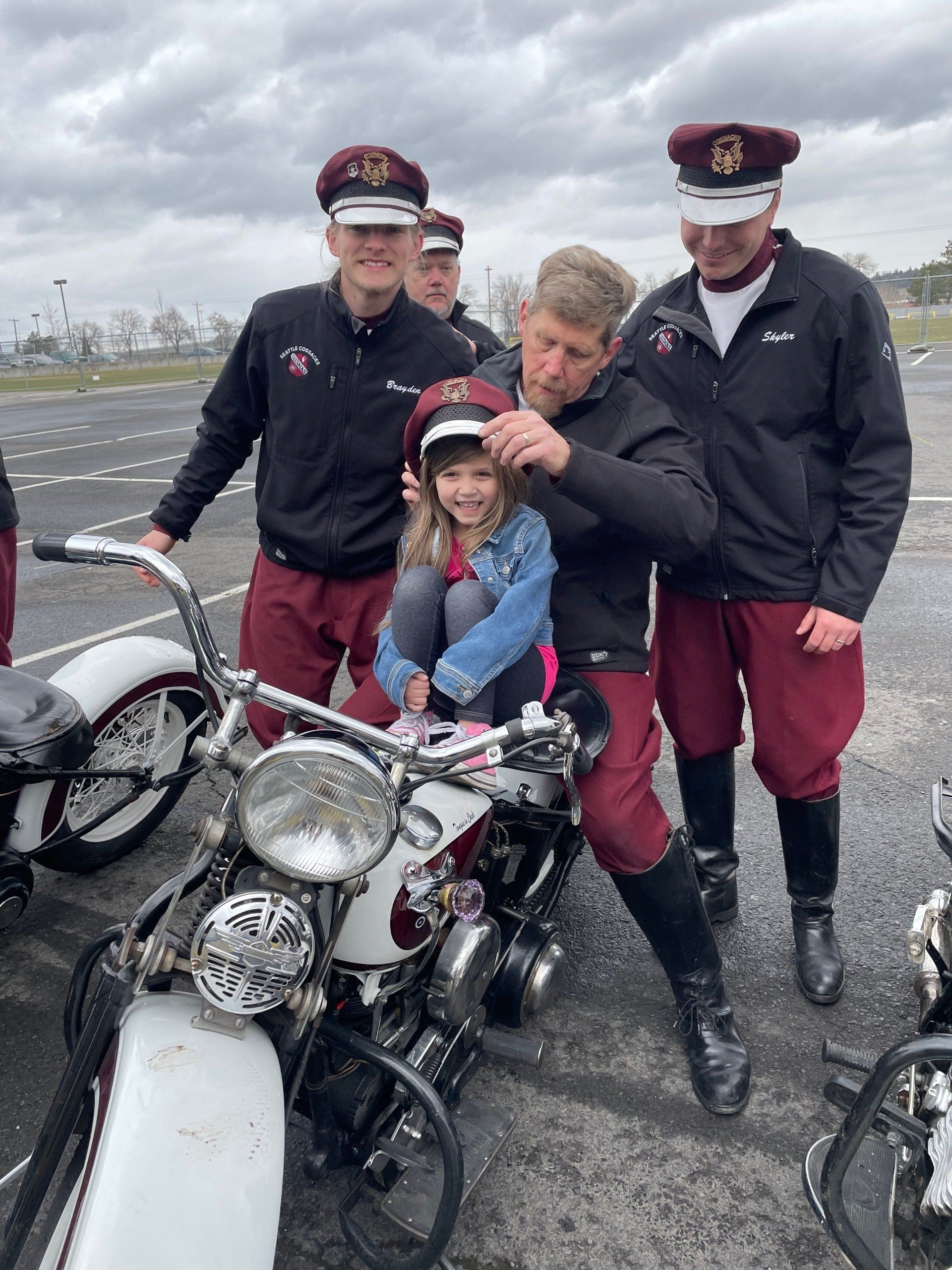 A group of men are standing around a motorcycle with a little girl on it.