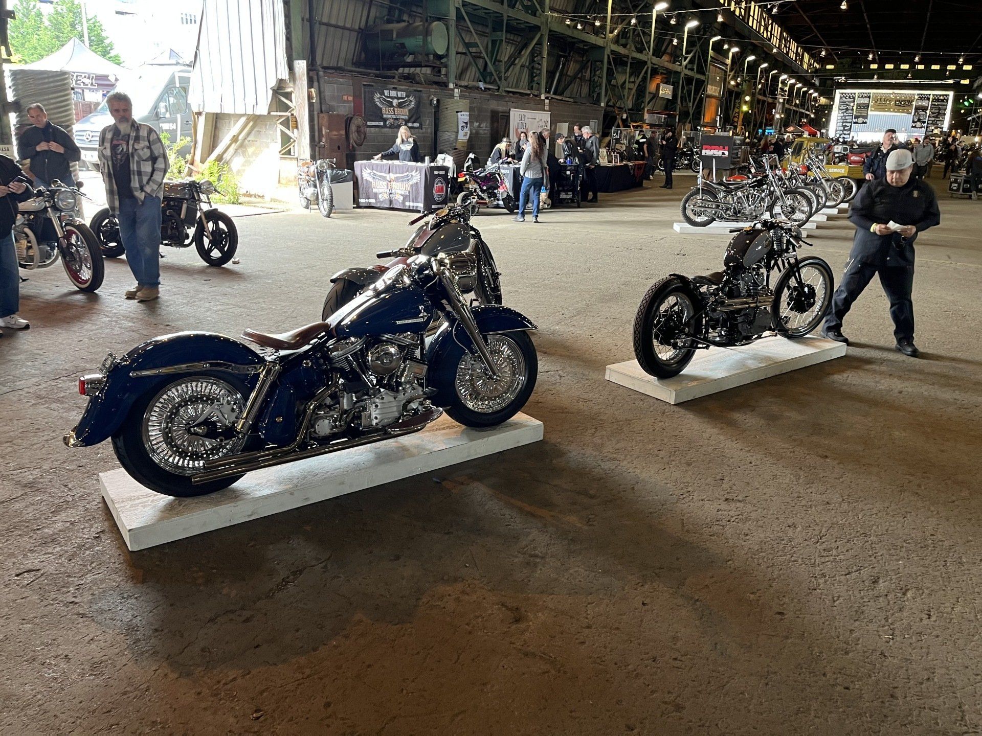 Two motorcycles are on display in a warehouse with people standing around them