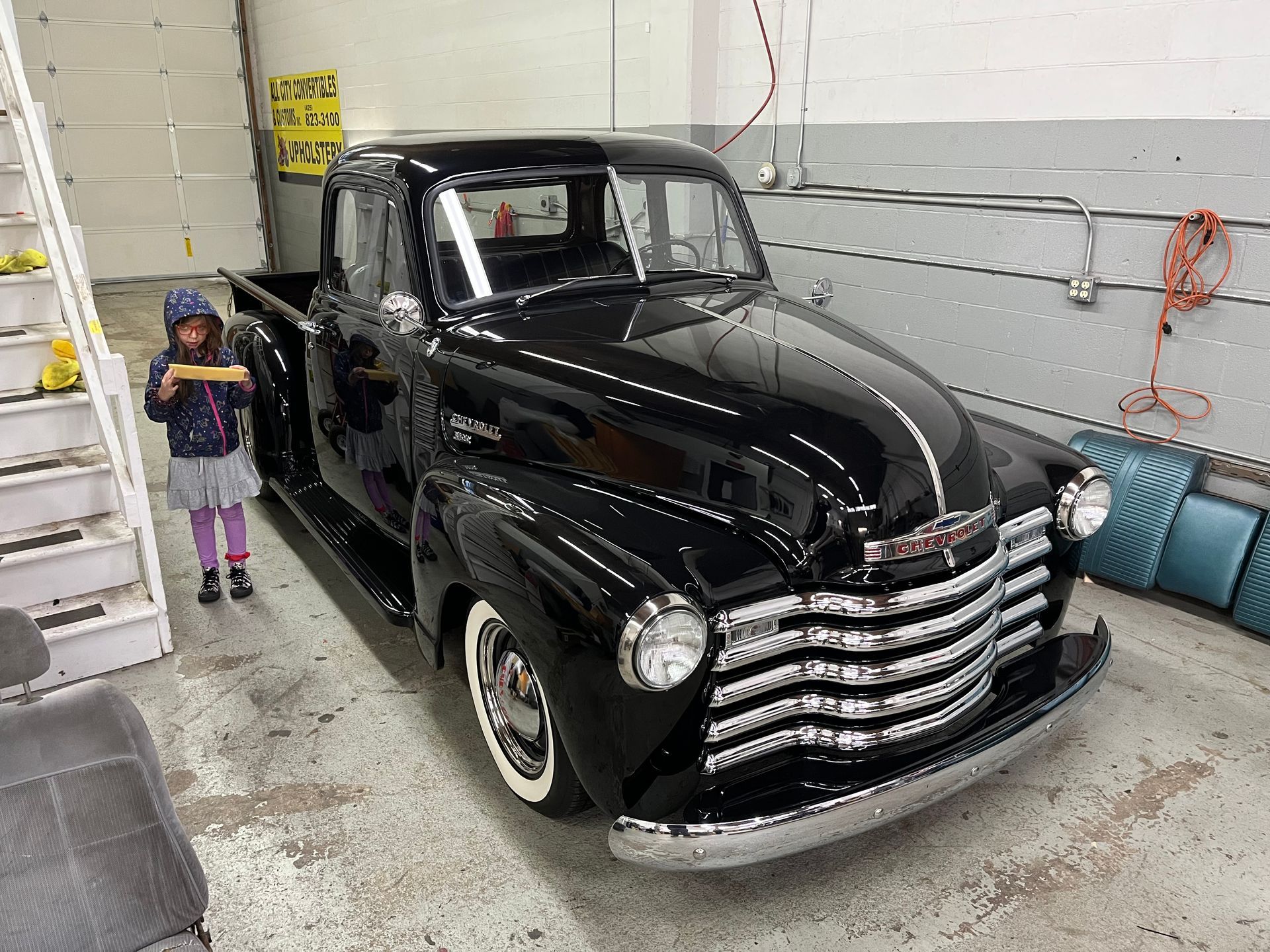 A little girl is standing next to an old black truck in a garage.