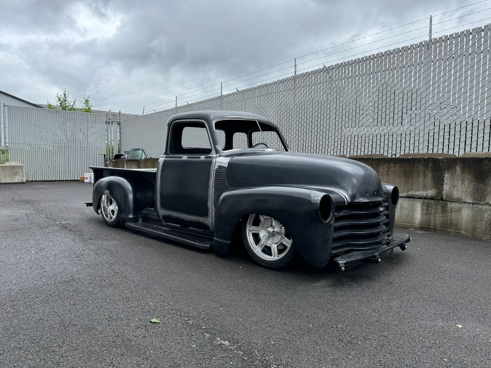 An old black truck is parked in a parking lot next to a fence.