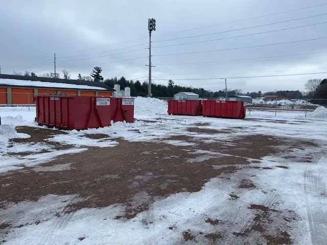 Two red dumpsters sit on a snowy, slushy gravel lot near storage buildings and a light pole under an overcast sky.
