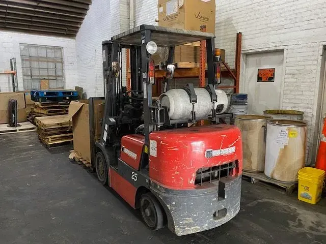 A red forklift parked in a warehouse, with a propane tank mounted on the back and pallets stacked to the side.