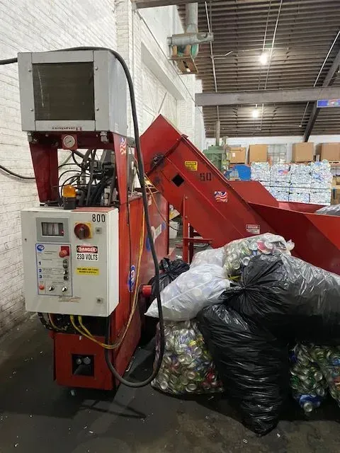A red industrial can baler machine in a warehouse, with bags of aluminum cans piled in front of it.