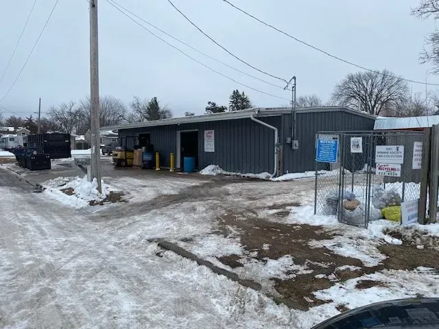 A grey warehouse building in a snowy area, featuring a forklift near the entrance and a metal cage with bagged items.
