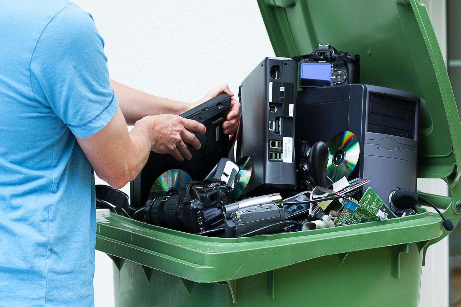 A person in a blue shirt places electronic waste, including computer towers and discs, into a green recycling bin.